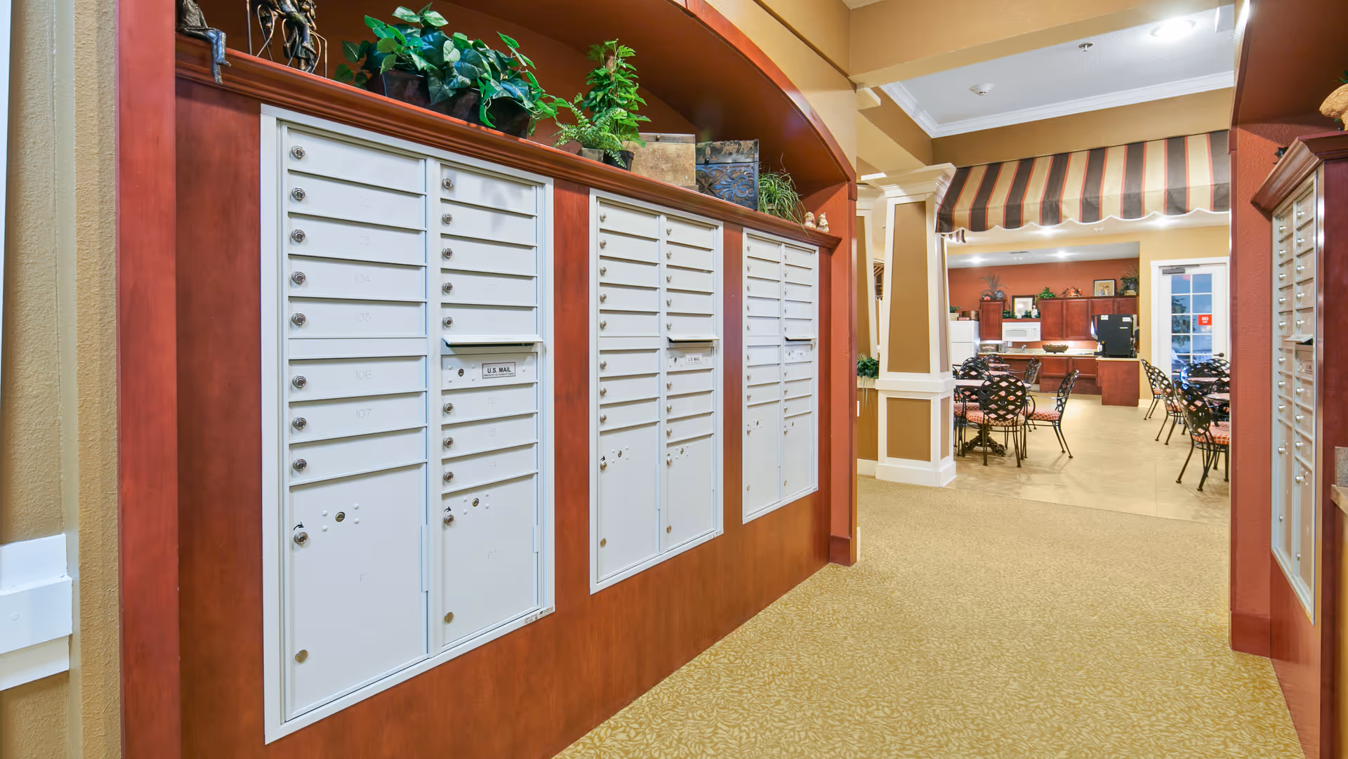 Mailboxes mounted on a wood-paneled wall in a facility hallway with a communal dining area and tables visible in the background.