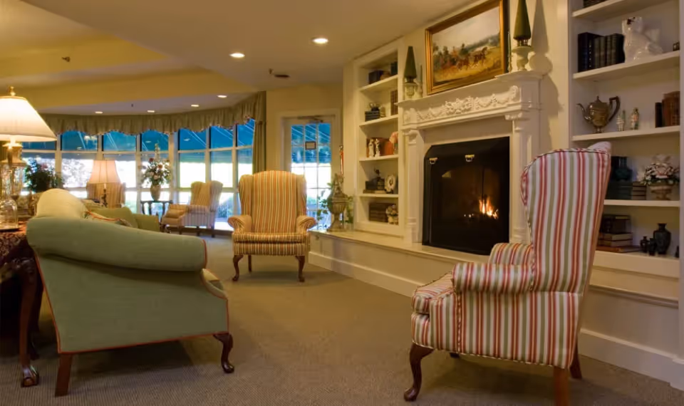 A cozy living room area with a lit fireplace surrounded by built-in shelves filled with books and decorative items. The room features several upholstered armchairs with striped patterns and a green sofa. Large windows with green awnings allow natural light to fill the space, and there are table lamps providing additional warm lighting.