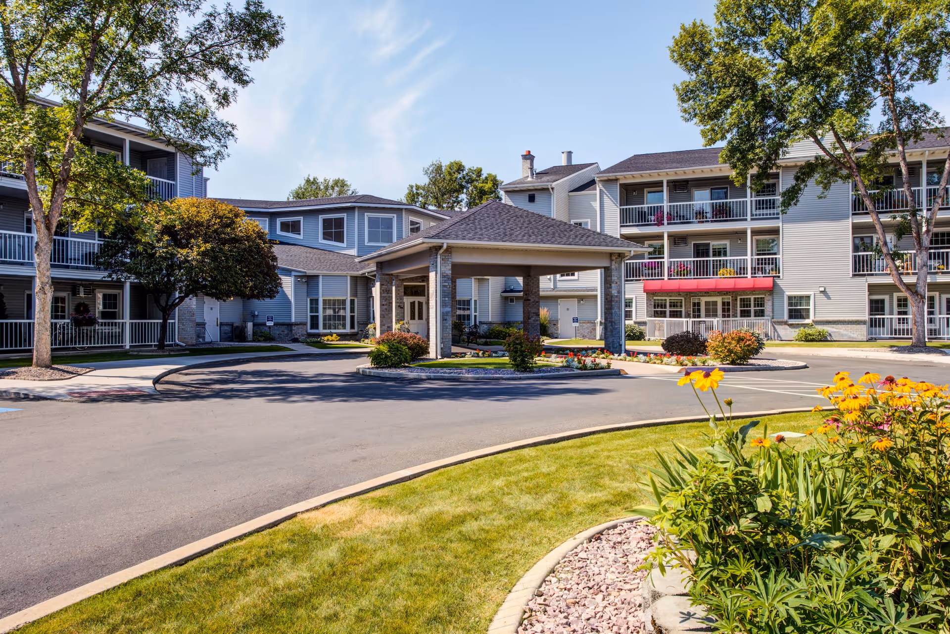 Exterior view of The Village Senior Residence showing a multi-story building with balconies, a covered entrance driveway, landscaped garden with flowers and trees, and a clear blue sky.