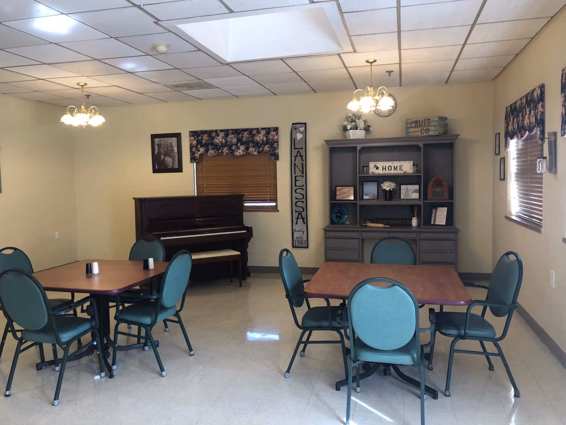 A common room with two square tables each surrounded by four green chairs. There is a piano against the far wall beneath a window with floral valance. A tall cabinet with shelves and drawers is on the right side, decorated with flowers, framed pictures, and signs including one that says 'HOME'. The walls are light yellow and the floor is tiled. Two ceiling light fixtures hang above the tables.