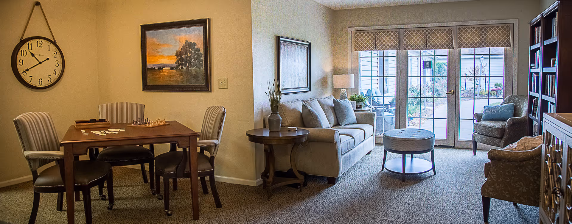 A cozy living room and dining area in a senior living facility. The dining area has a wooden table with four striped upholstered chairs, a chessboard, and a wall clock. The living room features a beige sofa, a round ottoman, two armchairs, a side table with a lamp, and a large window with glass doors leading outside. The room is decorated with framed artwork and a bookshelf.