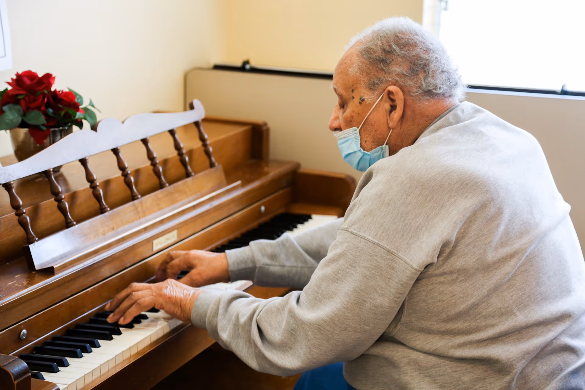 An elderly man wearing a face mask and a gray sweatshirt is playing a wooden piano in a well-lit room with a window. A pot of red flowers is placed on top of the piano.