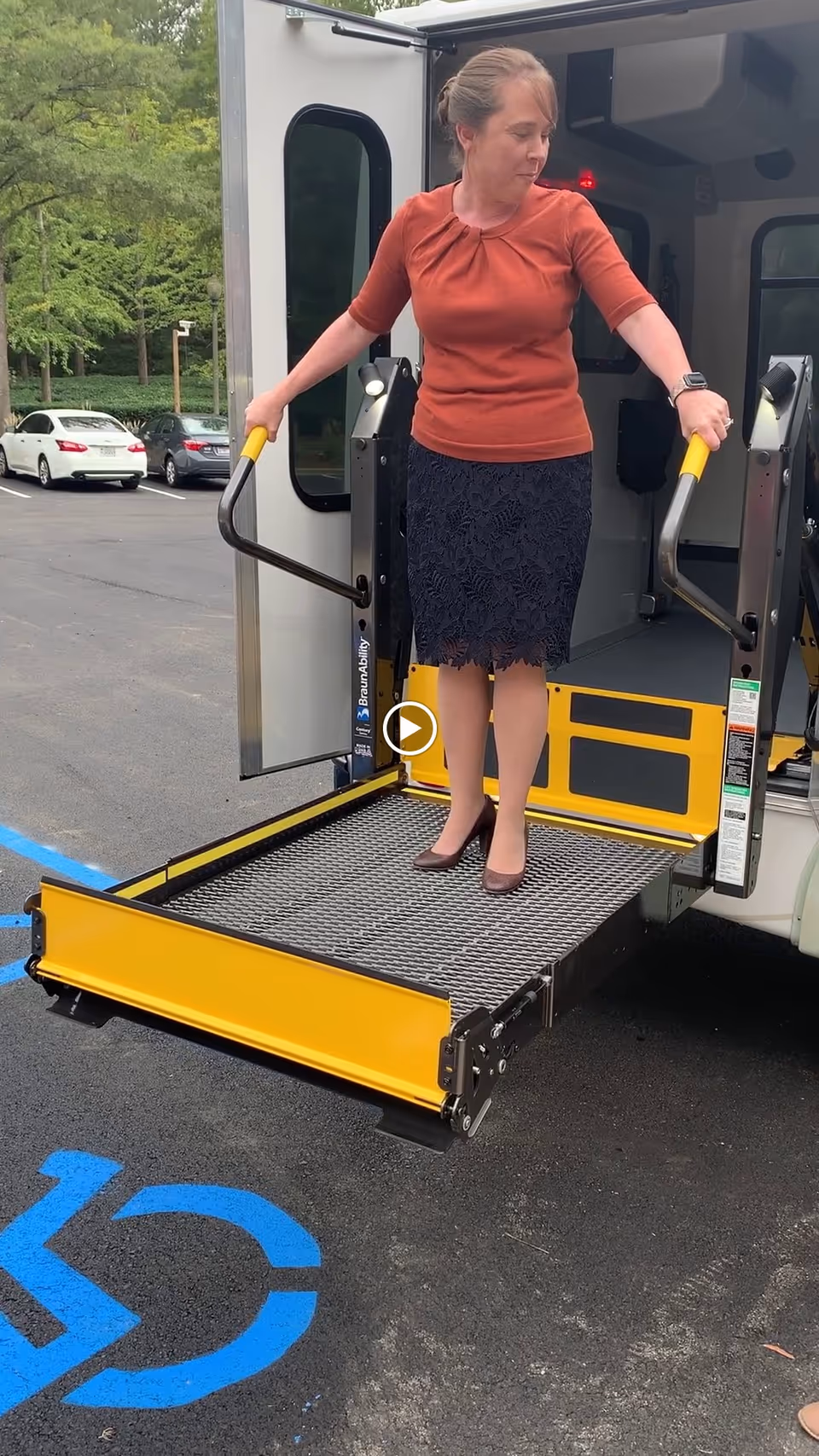 A woman in a rust-colored top and black skirt stands on a yellow wheelchair lift platform attached to a vehicle, holding onto the handrails. The vehicle door is open, and the lift is lowered to the ground in a parking lot with a blue handicap parking symbol visible on the asphalt.