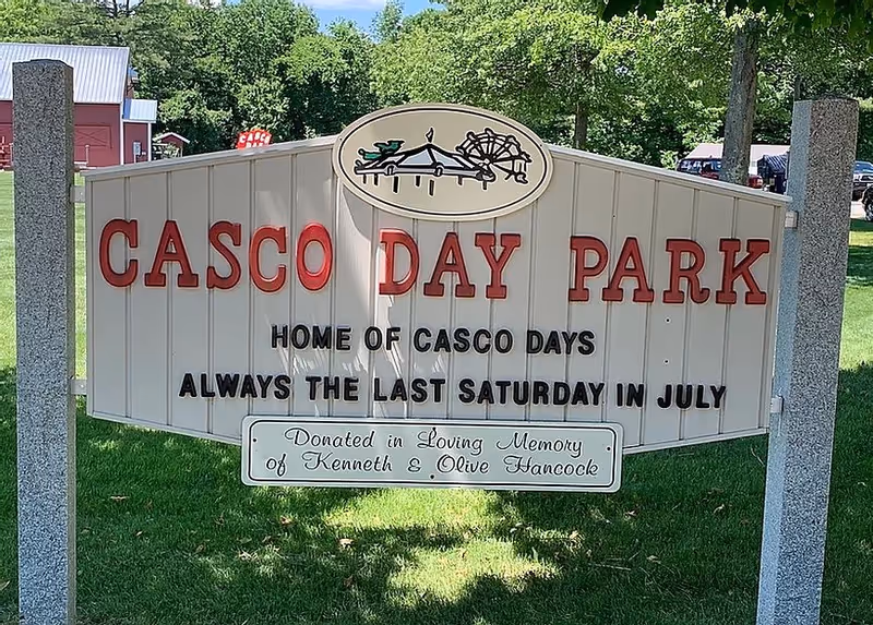 A sign for Casco Day Park with red and black lettering on a white background, supported by two stone posts. The sign reads: 'CASCO DAY PARK, HOME OF CASCO DAYS, ALWAYS THE LAST SATURDAY IN JULY.' There is a smaller plaque at the bottom that says, 'Donated in Loving Memory of Kenneth & Olive Hancock.' Trees and a red barn-like building are visible in the background.