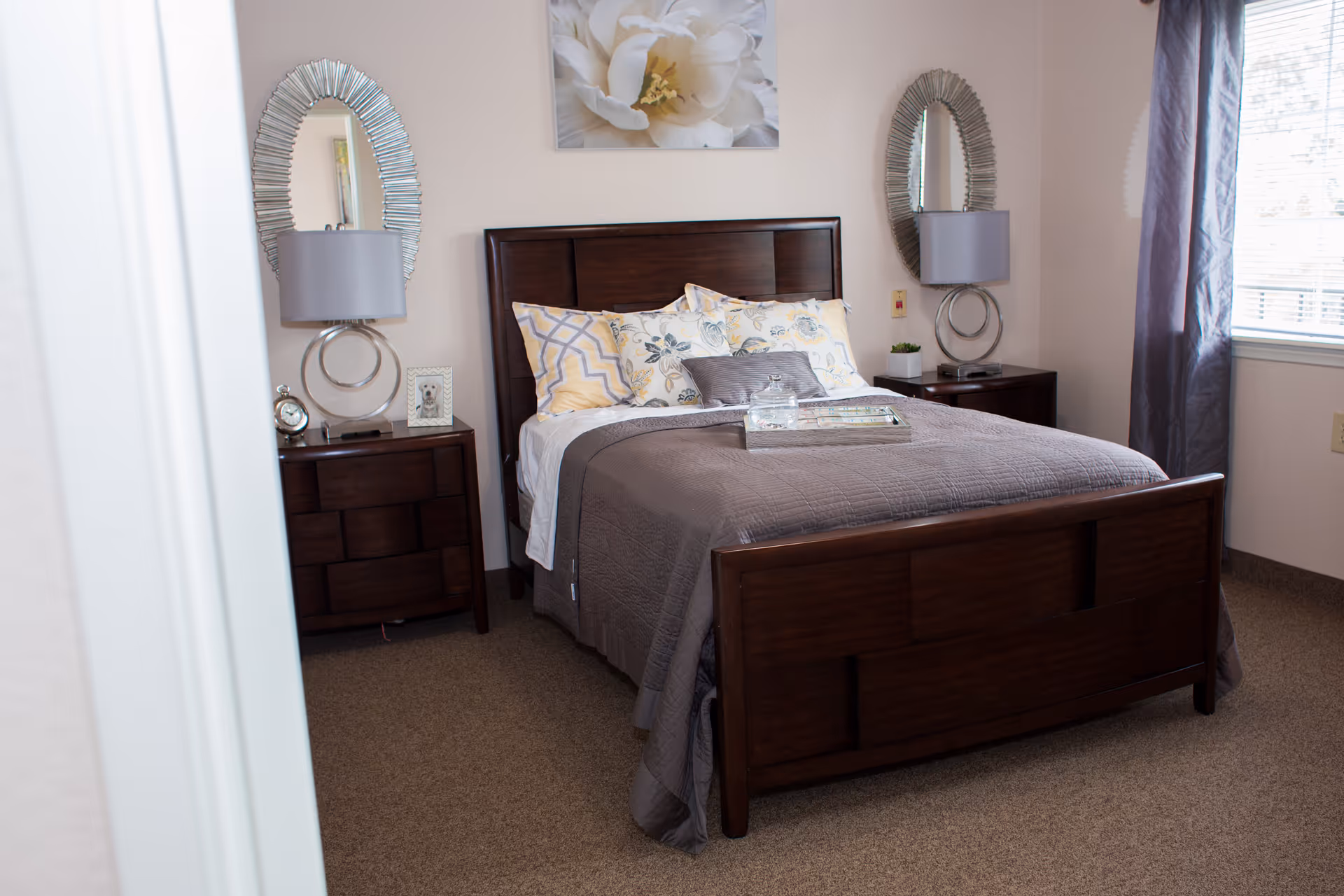 A neatly made bed with a dark wooden frame in a bedroom. The bed has multiple pillows with floral and geometric patterns and a gray quilt. On each side of the bed are matching dark wooden nightstands with modern circular lamps and decorative items. Two oval mirrors hang on the wall above the nightstands, and a large floral artwork is centered above the headboard. A window with gray curtains is on the right side of the room.