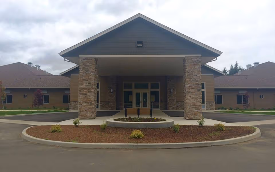 Front entrance of a single-story senior living facility with a covered porte-cochère, stone columns, and a circular driveway planter.