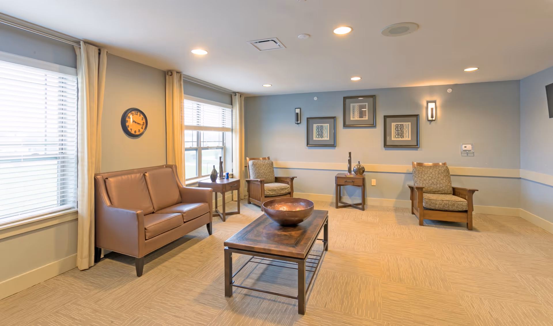 A well-lit living room with beige carpet, light blue walls, and recessed ceiling lights. The room features a brown leather loveseat, two wooden armchairs with patterned cushions, a wooden coffee table with a decorative bowl, two side tables with vases, a wall clock, framed artwork, and large windows with beige curtains and blinds.