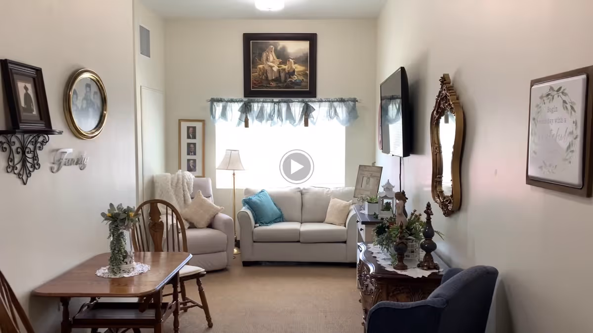 A cozy living room with a beige sofa and armchair adorned with cushions, a wooden dining table with chairs, decorative wall art including family photos and framed quotes, a flat-screen TV mounted on the wall, and a wooden sideboard with plants and figurines. A window with sheer blue curtains lets in natural light.
