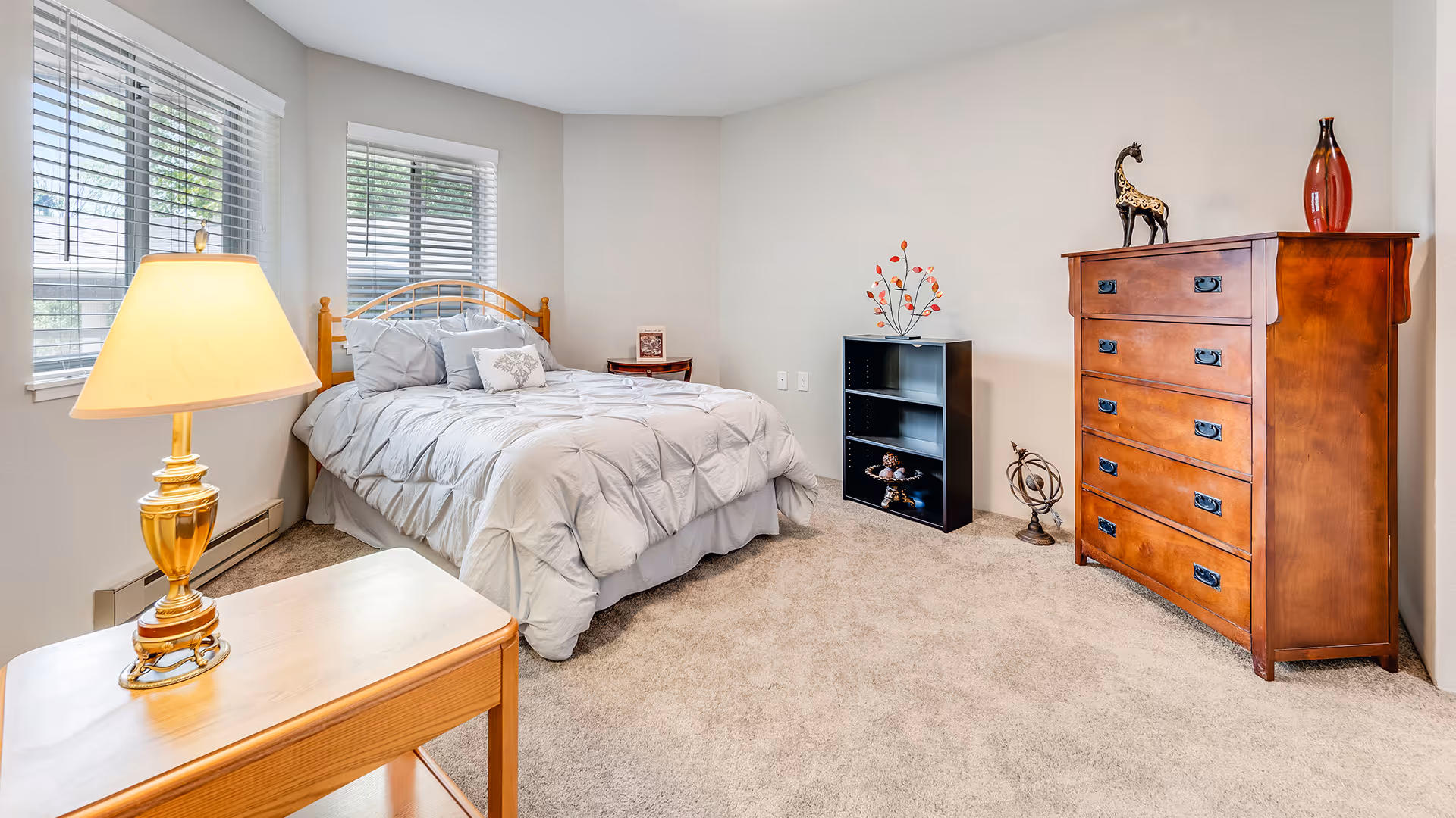 A bedroom with a neatly made bed featuring light gray bedding, two windows with white blinds, a wooden nightstand with a lamp, a wooden dresser with decorative items on top, and a small black bookshelf with ornaments. The room has beige carpet and light-colored walls.