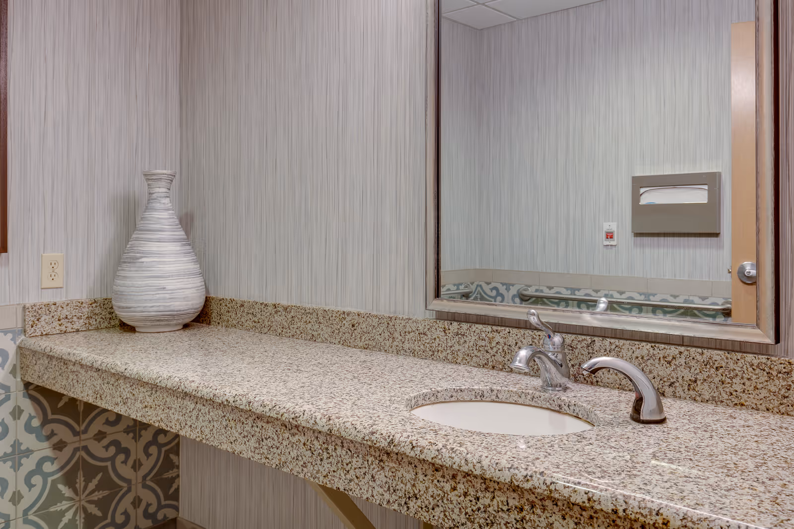 A bathroom countertop with a built-in sink and two silver faucets. There is a large decorative vase on the left side of the granite countertop. A large mirror is mounted on the wall above the sink, reflecting a paper towel dispenser and a wall-mounted emergency button. The wall has a textured light gray wallpaper, and the lower part of the wall features patterned tiles.