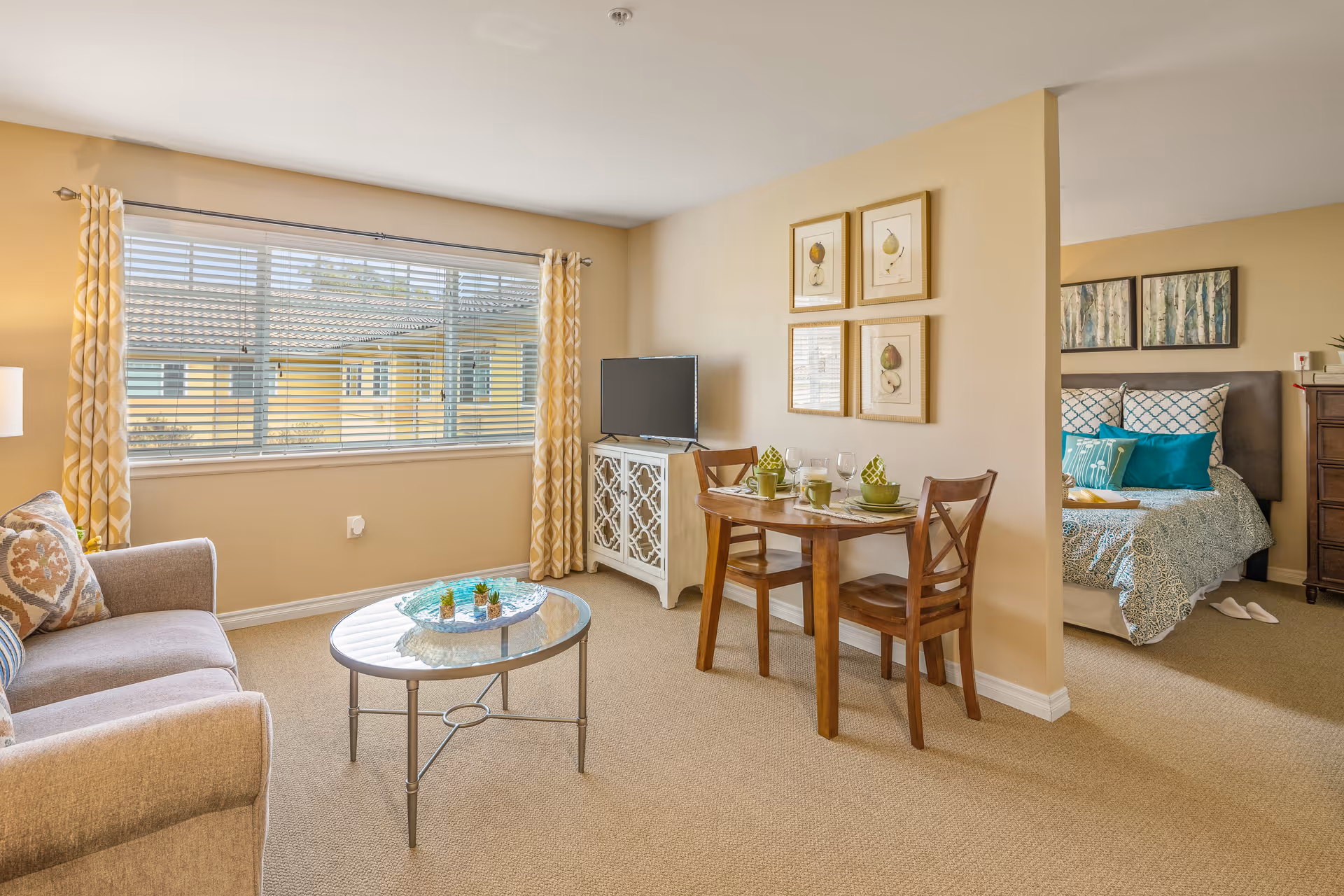 Well-lit living area with a sofa, coffee table, small dining table, TV and an adjoining open bedroom visible through a partial wall.