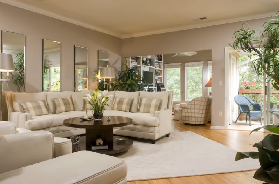 Bright living room with a beige sectional sofa, round coffee table, wall mirrors and a sliding door opening to a deck.