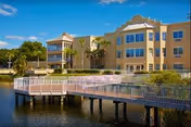 Exterior view of a multi-story beige building with large windows and balconies, surrounded by palm trees and greenery, adjacent to a body of water with a wooden boardwalk extending over the water under a clear blue sky.