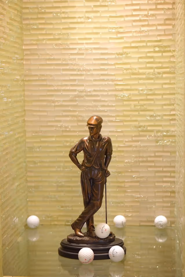 Bronze golfer statue on a glass shelf surrounded by golf balls against a light green textured tiled wall.