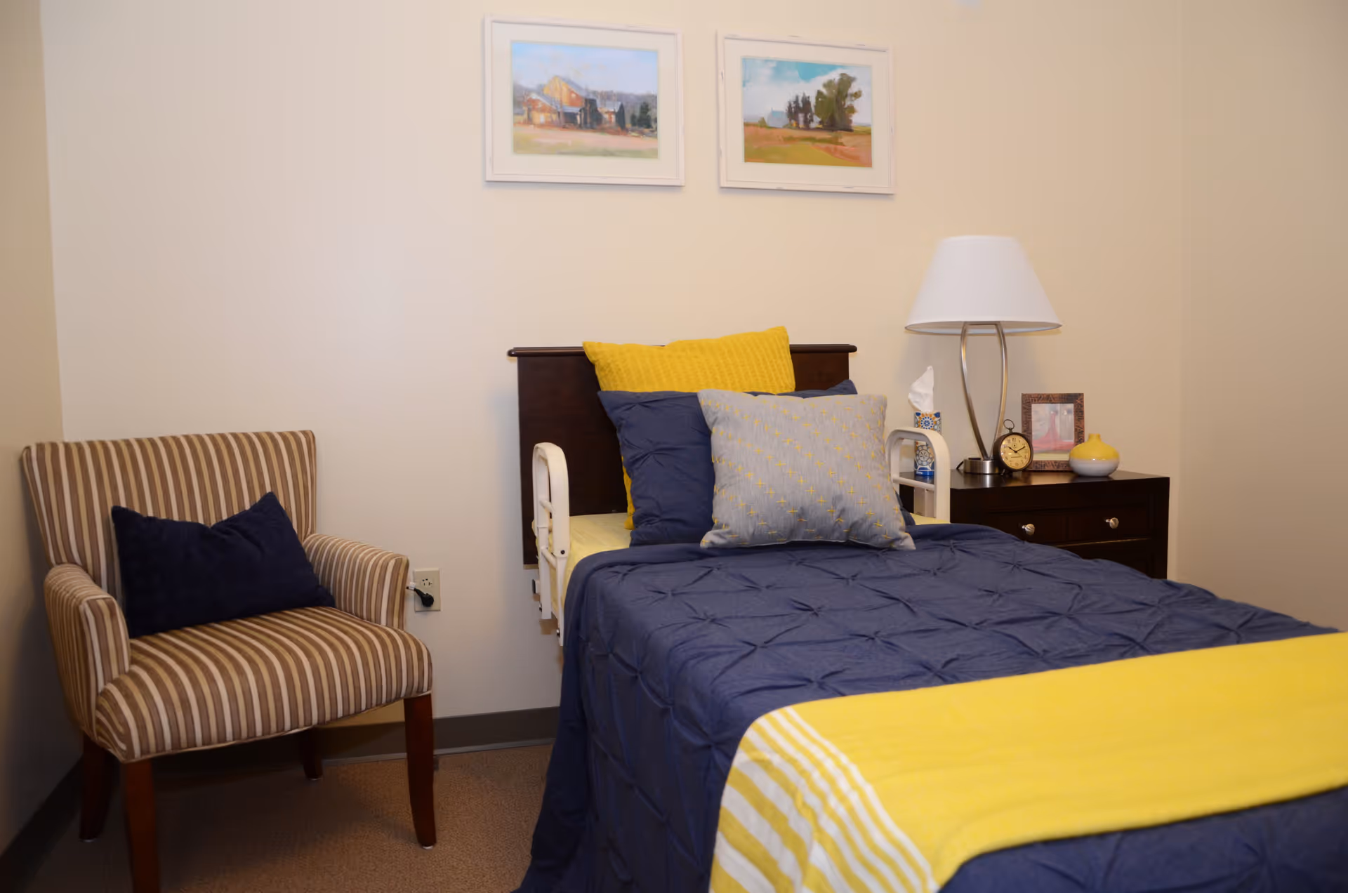 A bedroom in a senior living facility featuring a single bed with a dark blue comforter, yellow and gray pillows, and a yellow throw blanket. Next to the bed is a dark wooden nightstand with a lamp, a clock, a tissue box, and a small decorative item. A striped armchair with a dark blue pillow is positioned to the left of the bed. Two framed landscape paintings hang on the wall above the bed.