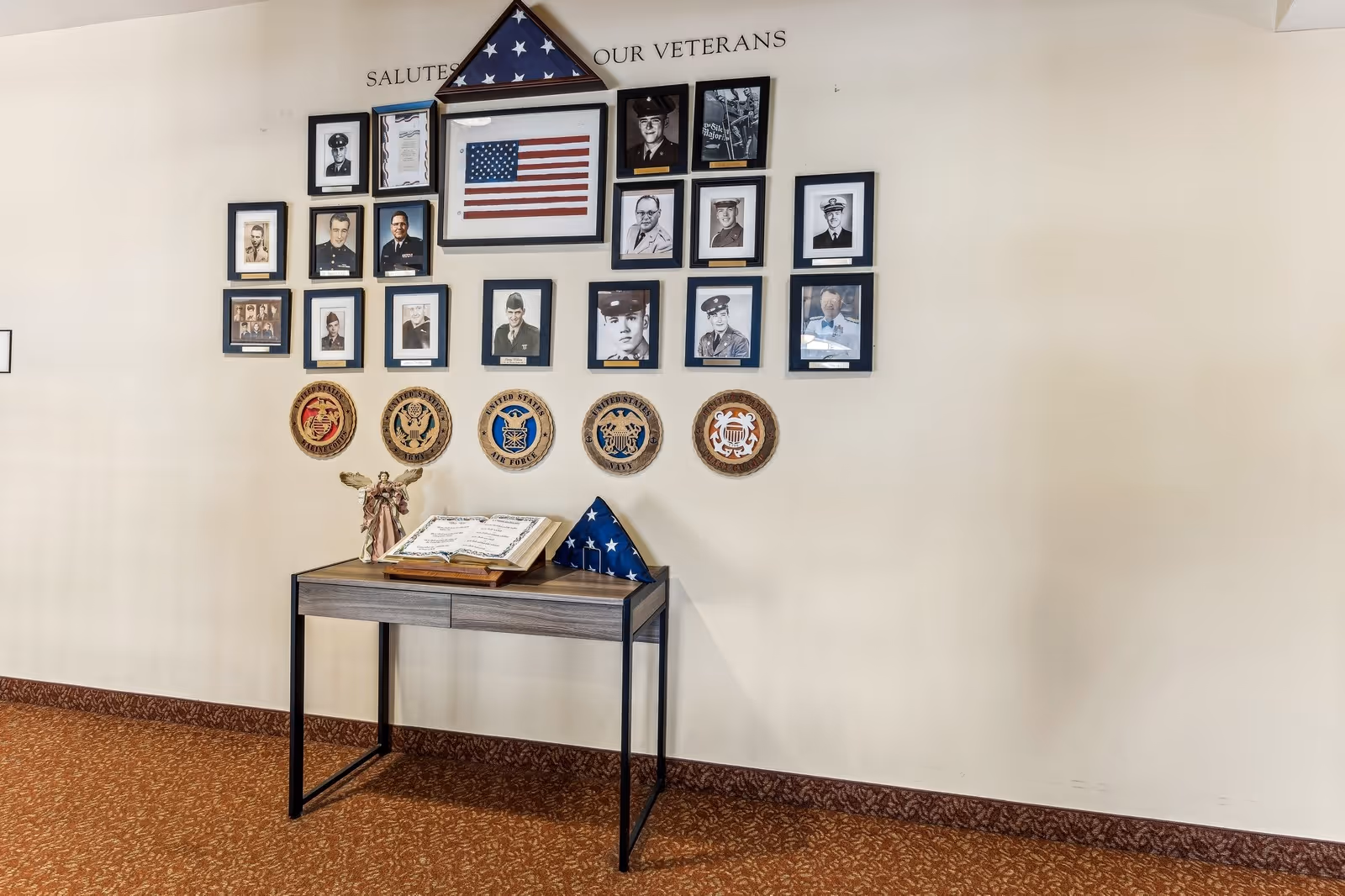 A memorial wall display honoring veterans with framed photographs of servicemen and servicewomen, an American flag, military insignias, and a folded flag on a small table beneath the display. The wall has the text 'Salutes Our Veterans' above the photos.