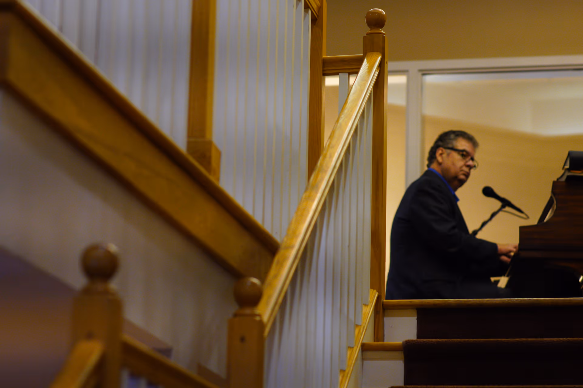 A man in a dark suit playing a piano with a microphone in front of him, viewed from the side of a wooden staircase with white balusters inside a building.