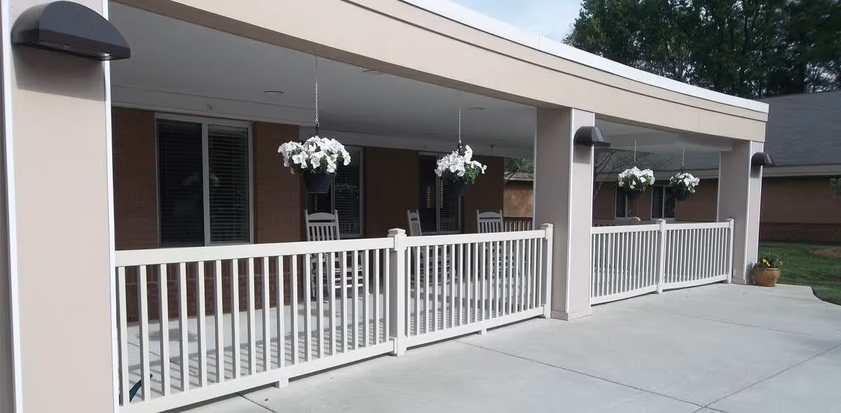 Covered outdoor patio area with white railings, hanging flower pots with white flowers, and several white rocking chairs. The patio is attached to a brick building with windows and is surrounded by a concrete walkway and green grass.