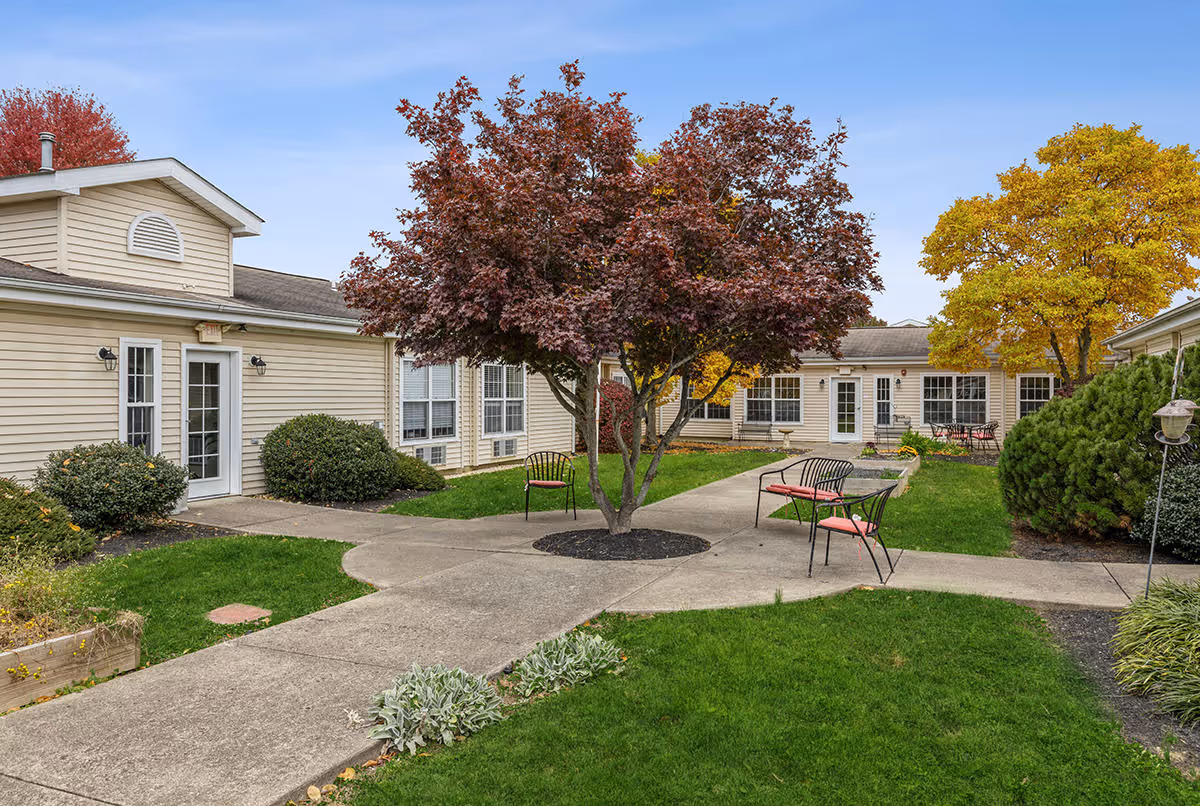 Outdoor courtyard area at Clay Gardens Place featuring a paved walkway, green grass, a tree with red leaves in the center, benches with red cushions, and beige buildings with multiple windows surrounding the courtyard. Trees with autumn foliage are visible in the background under a clear blue sky.