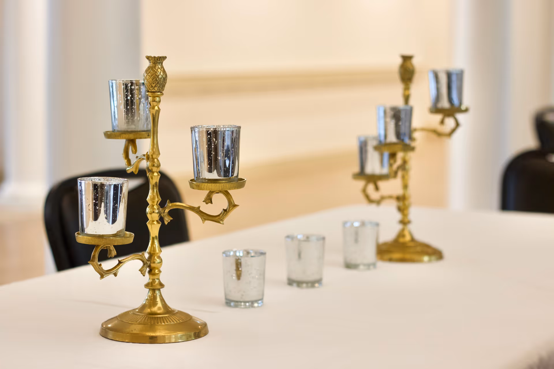 Brass candelabras and small glass votive holders arranged on a white-clothed table in an indoor event/dining space.