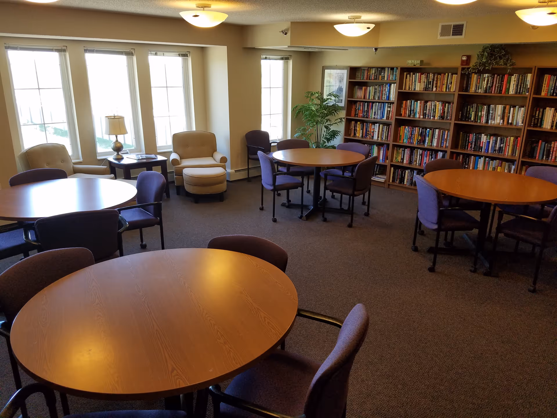 Bright senior common room with round wooden tables and chairs, bookshelves along one wall, and armchairs by windows.