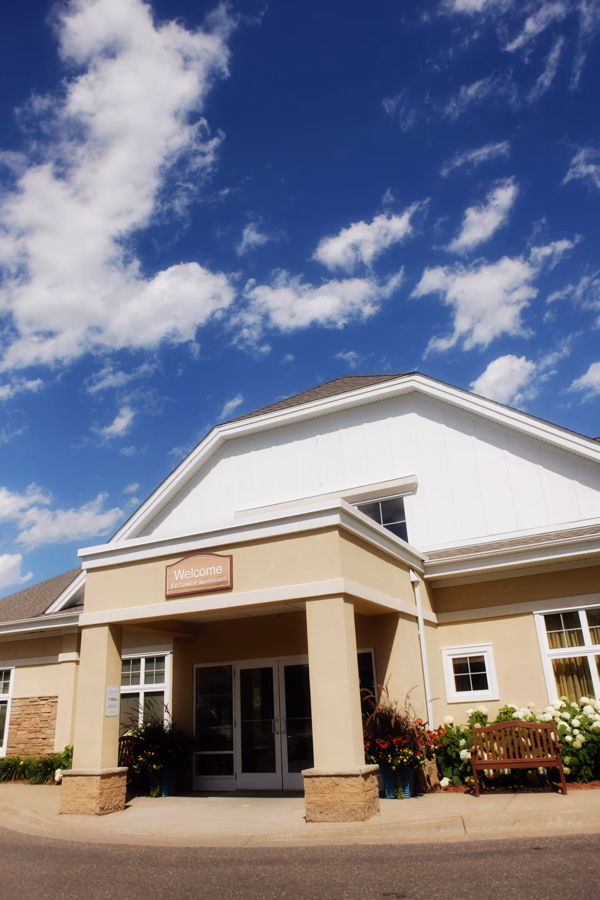 Front entrance of a senior living facility with a covered portico, welcome sign, bench and potted plants under a blue sky.