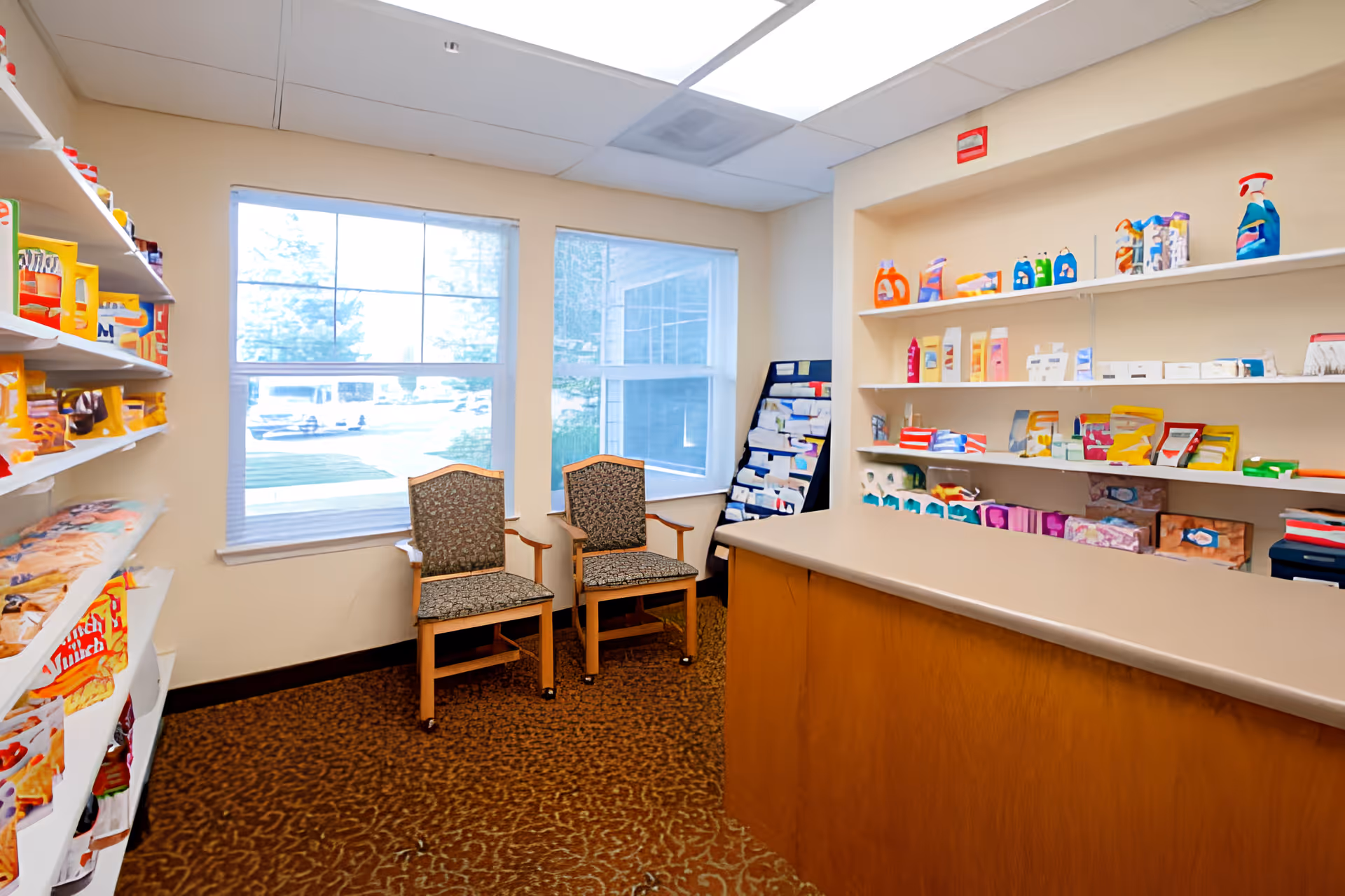 Small sundries/shop room with stocked shelves, a service counter, and two chairs by a window.