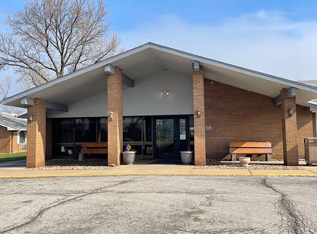 Front entrance of a brick senior living facility featuring a covered peaked portico with benches, potted plants, and glass double doors.
