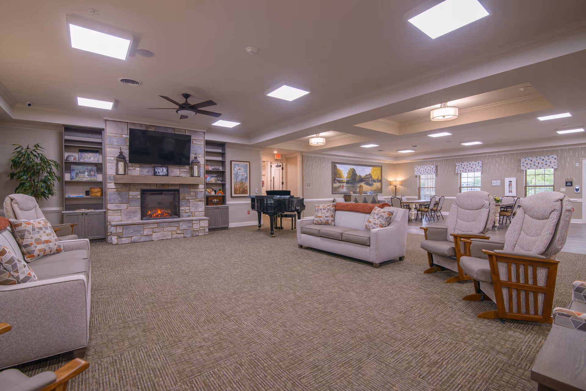 A spacious and well-lit living room area in a senior living facility featuring comfortable seating including sofas and armchairs arranged around a stone fireplace with a mounted flat-screen TV above it. The room has built-in shelves with decorative items, a ceiling fan, and a grand piano in the corner. In the background, there are dining tables and chairs near windows with patterned valances, and soft lighting fixtures on the ceiling.