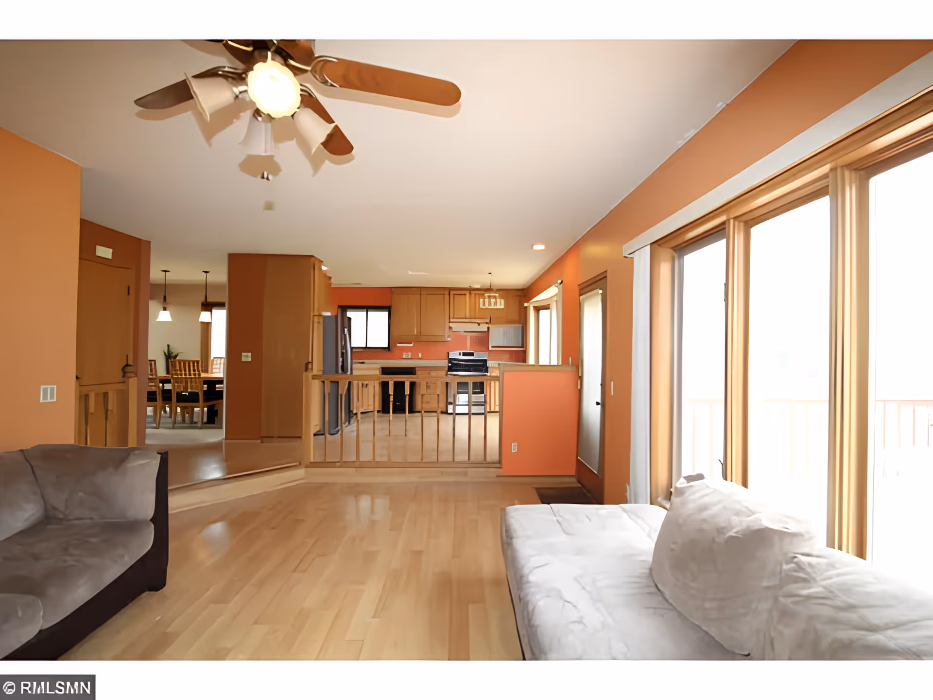 Interior view of a living room with light wooden flooring, two sofas, and large windows letting in natural light. The room opens up to a kitchen area with wooden cabinets, stainless steel appliances, and a dining area with a wooden table and chairs visible in the background. The walls are painted in a warm orange tone, and a ceiling fan with lights is mounted on the ceiling.