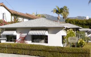 White single-story building with striped awnings, hedges, and palm trees in front.