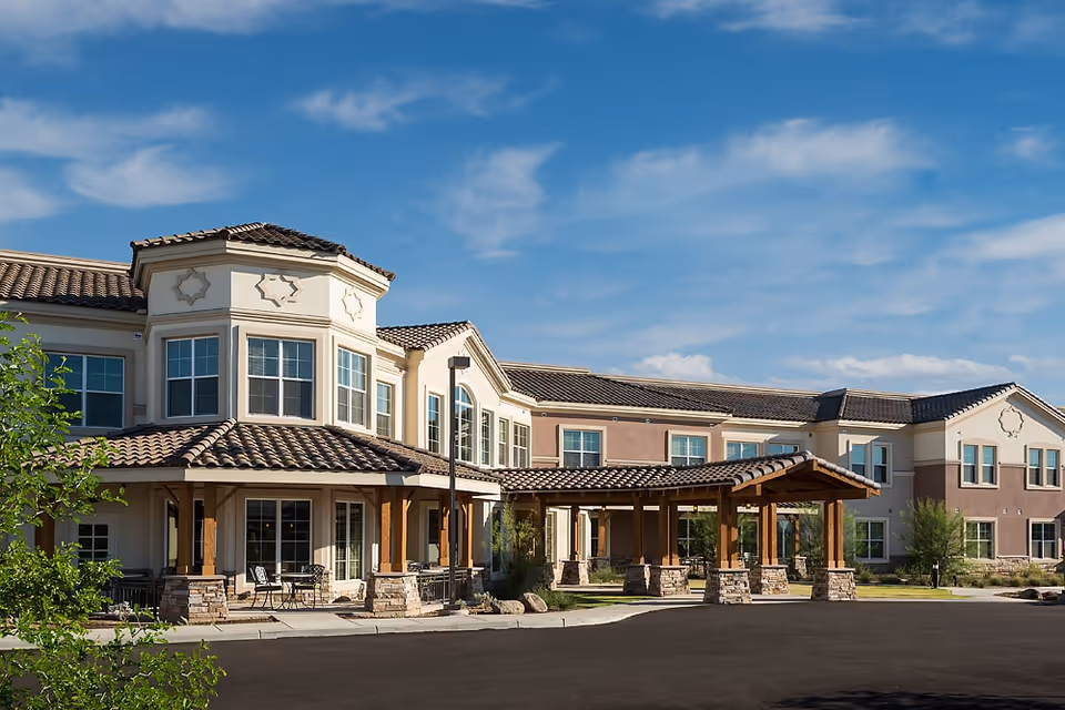Exterior view of MorningStar At Arrowhead senior living facility showing a two-story building with beige and brown walls, multiple windows, a covered entrance with wooden beams and stone pillars, and a clear blue sky with some clouds.