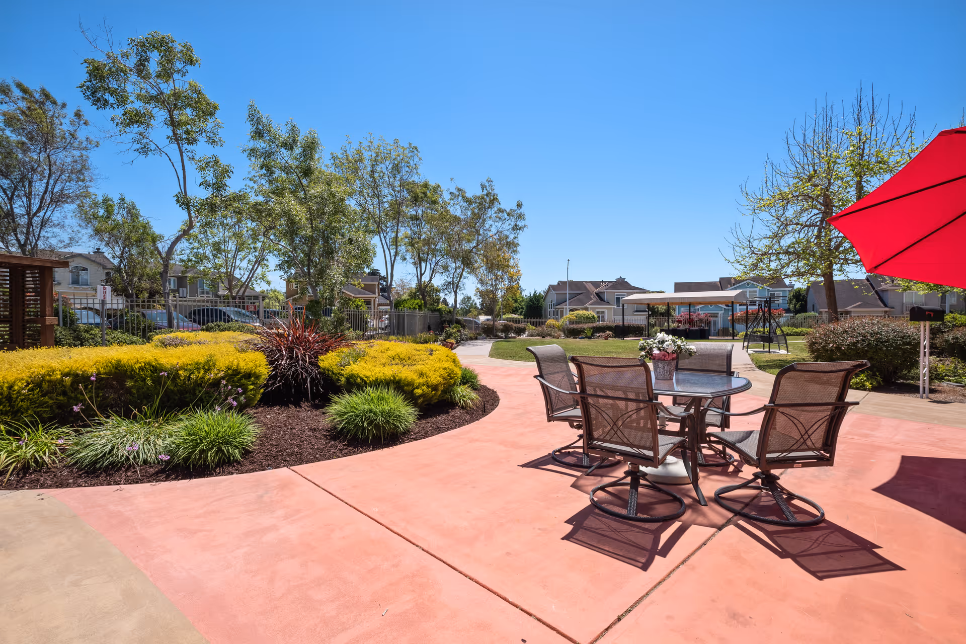 Outdoor patio area with a round glass table and four metal mesh chairs under a red umbrella. The patio is surrounded by landscaped greenery, including bushes, small trees, and flowering plants, with residential buildings visible in the background under a clear blue sky.