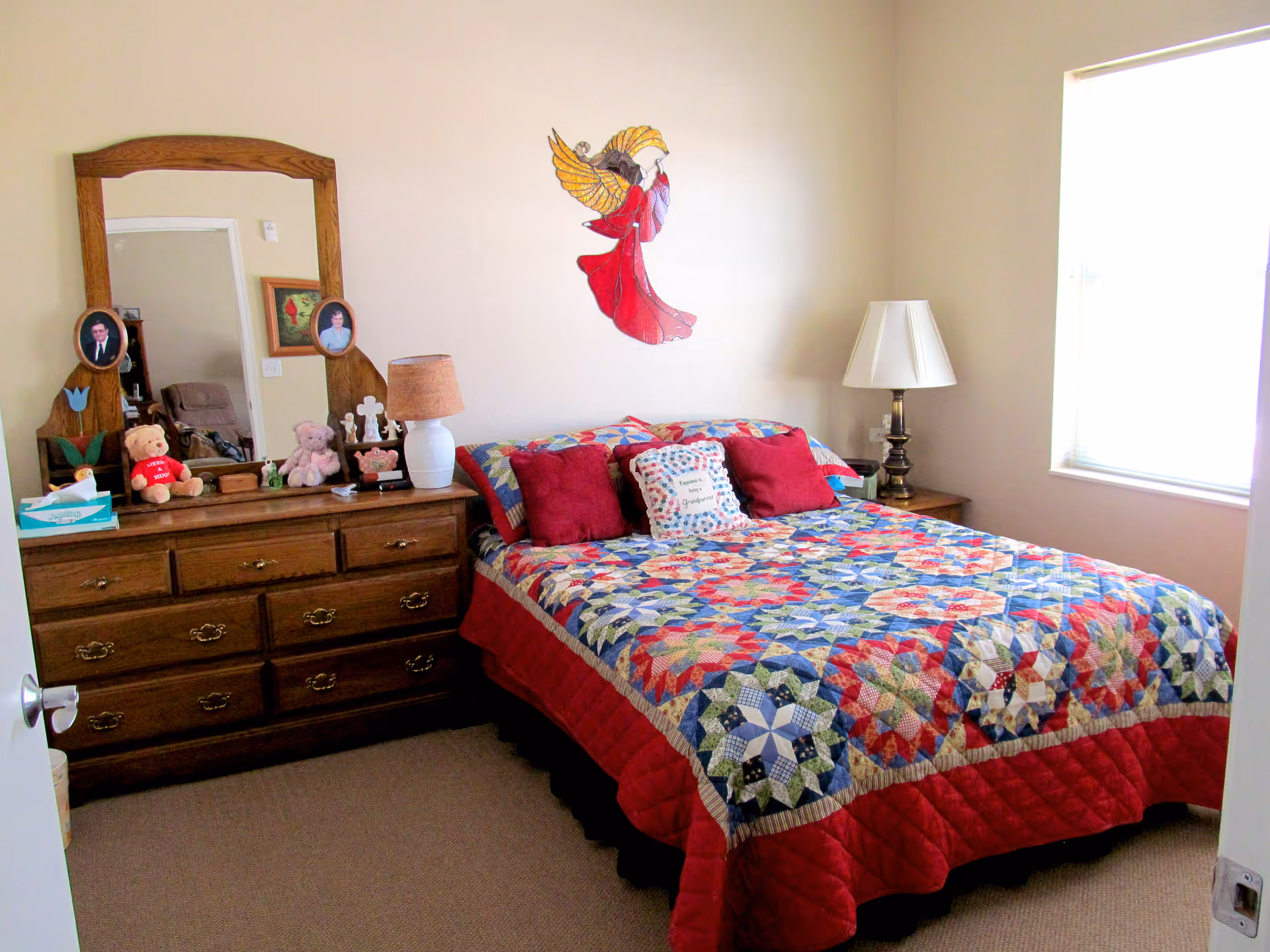 A cozy bedroom with a colorful patchwork quilt on the bed, two red pillows, and a decorative pillow that says 'Happiness is a Grandparent'. There is a wooden dresser with a large mirror, two framed photos, teddy bears, and a lamp. A stained glass angel decoration hangs on the wall above the bed. A window with blinds allows natural light into the room.