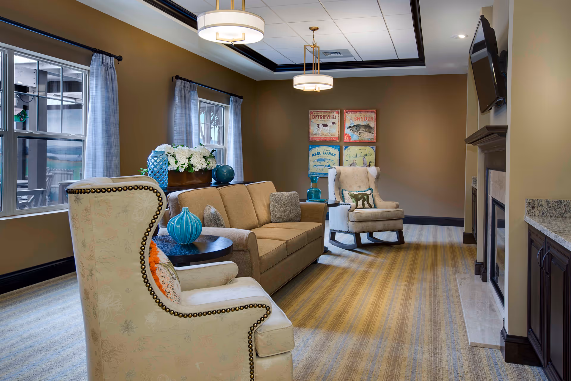 A cozy living room in Harbor at Harmony Crossing featuring a beige sofa with cushions, two upholstered armchairs with decorative pillows, a fireplace with a mounted TV above it, and a side table with blue decorative vases. The room has large windows with light curtains, a striped carpet, and framed vintage-style artwork on the wall.