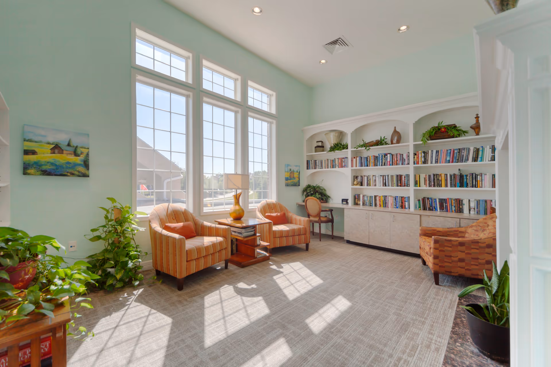 A bright and cozy living room area with large windows letting in natural light. The room features two striped armchairs with orange cushions, a small wooden side table with a lamp, a built-in white bookshelf filled with books and decorative plants, and additional seating including an orange patterned armchair and a wooden chair at a small desk. There are several green plants and a painting on the light blue walls.