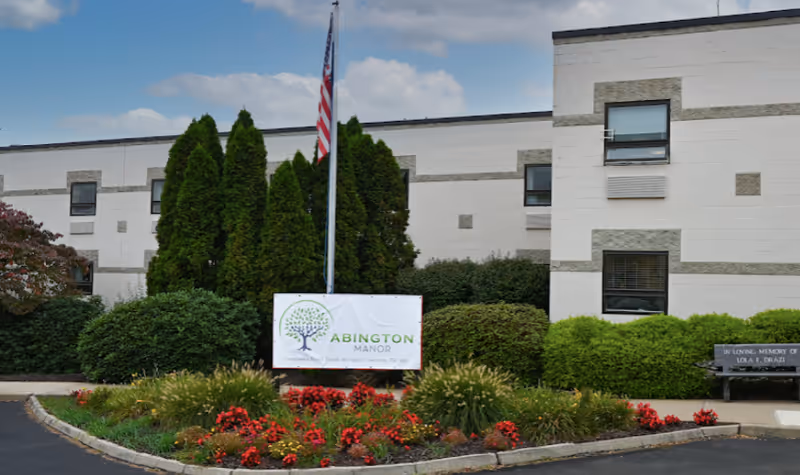 Exterior view of Abington Manor building with a white sign displaying the facility name in front of tall green bushes and a flower bed with red flowers. An American flag is on a flagpole behind the sign, and the building has white walls with rectangular windows.