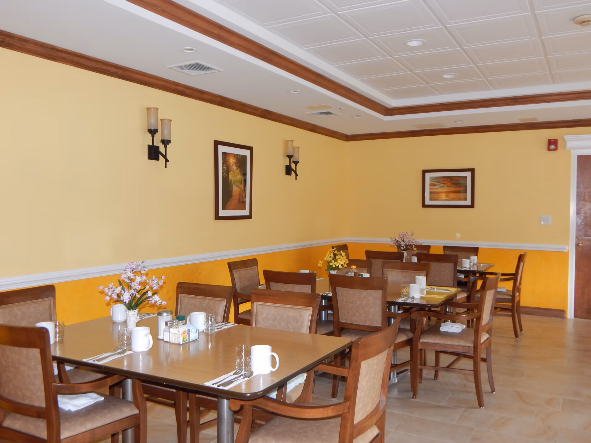 A dining room with several wooden tables and chairs arranged neatly. Each table is set with white mugs, glasses, napkins, and silverware. There are small flower arrangements on some tables. The walls are painted yellow with white trim and decorated with framed pictures and wall sconces. The ceiling has a recessed design with lighting fixtures.