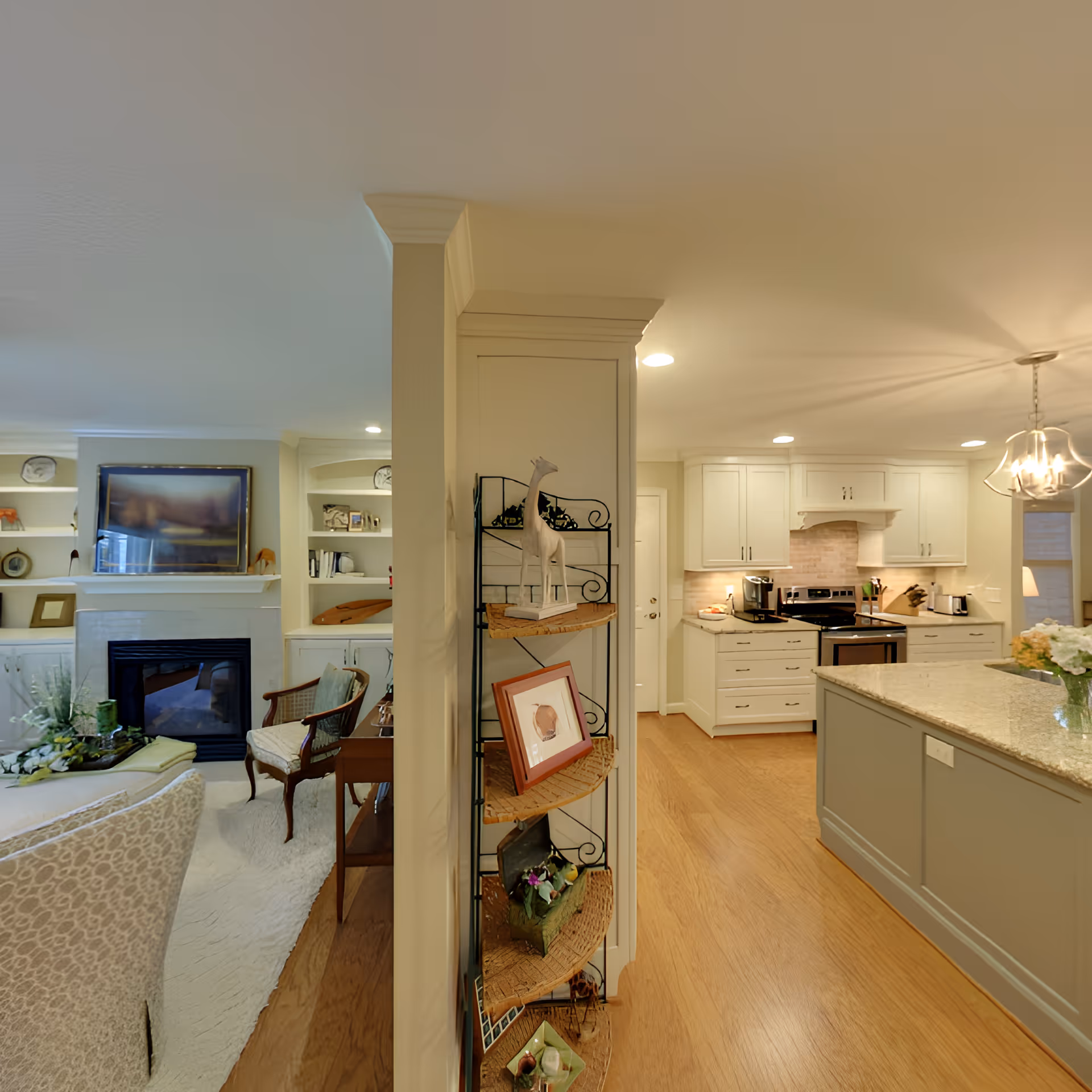Interior view of a senior living facility showing a cozy living room with a fireplace, armchair, and built-in shelves on the left, and a modern kitchen with white cabinets, stainless steel appliances, and a large island with a granite countertop on the right. A decorative corner shelf with various items is positioned in the center.