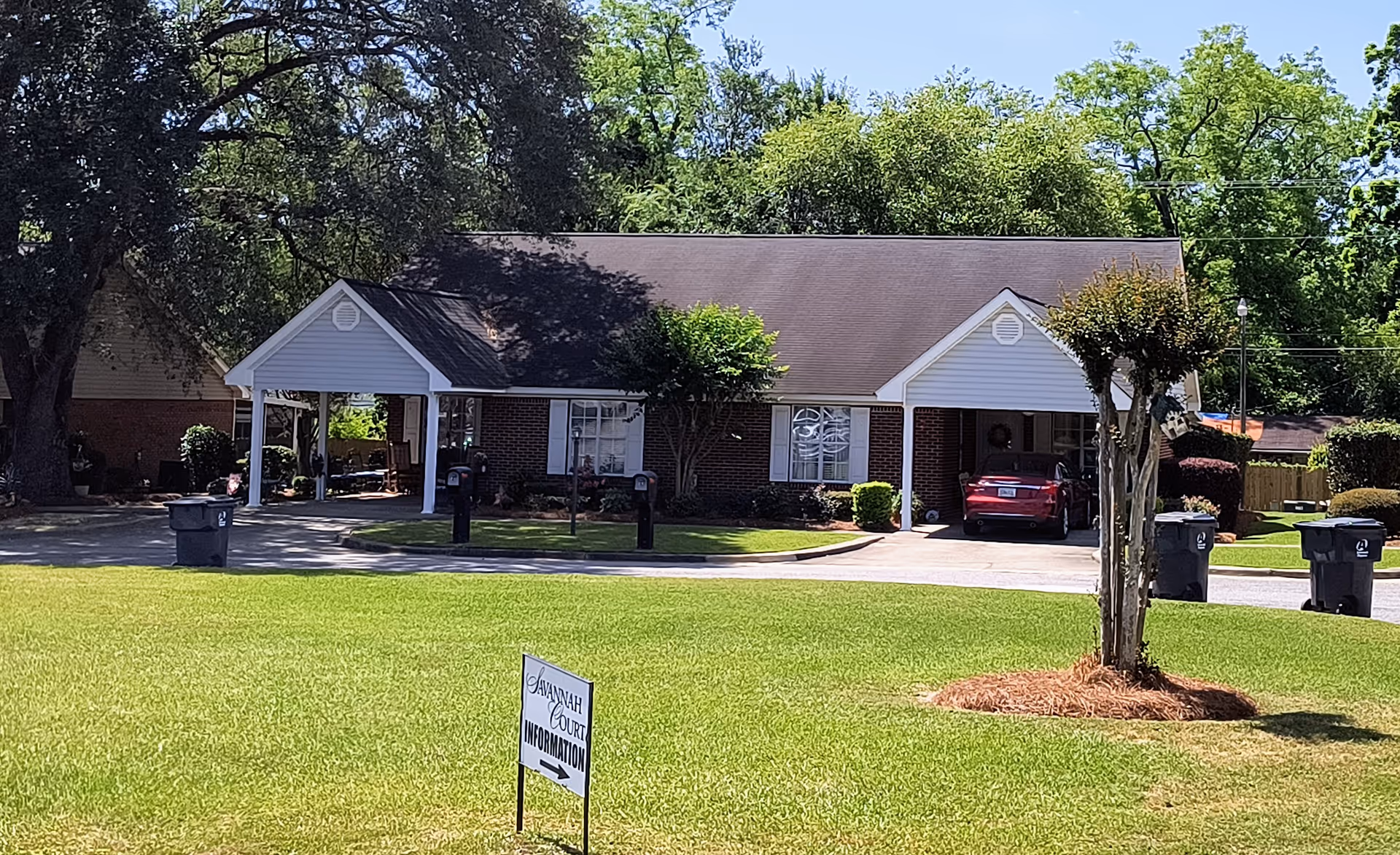 Exterior view of a single-story brick building with a gable roof, white trim, and a covered carport. There is a red car parked under the carport, several trash bins along the driveway, and a small tree with mulch around its base in the foreground. A sign on the lawn reads 'Savannah Court Information' with an arrow pointing to the right. The area is surrounded by green grass and trees under a clear blue sky.