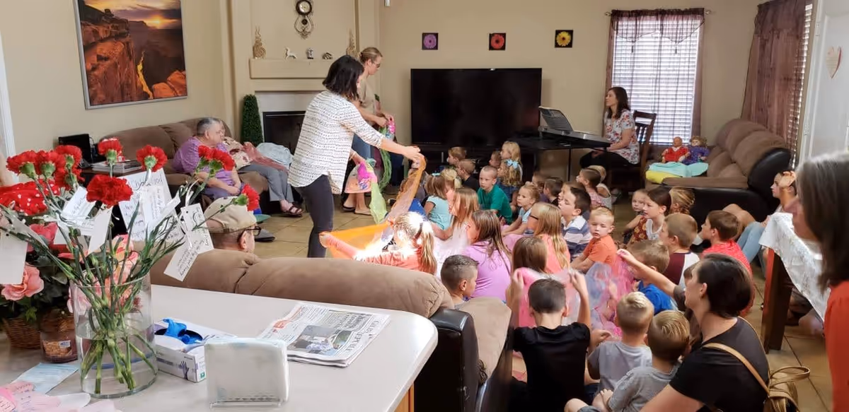 A group of children sitting on the floor in a living room, attentively watching a woman who is standing and holding colorful scarves. Several elderly people are seated on couches around the room. A woman is playing a keyboard in the background. The room has a large TV, a fireplace, and windows with curtains. A vase with red flowers and notes is on a nearby counter.