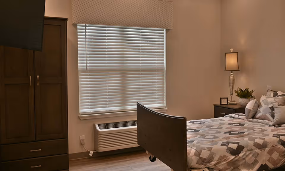 A cozy bedroom in a skilled nursing facility featuring a bed with patterned bedding, a wooden wardrobe, a nightstand with a lamp, a small clock, and a plant. A window with closed blinds and an air conditioning unit below it are also visible.