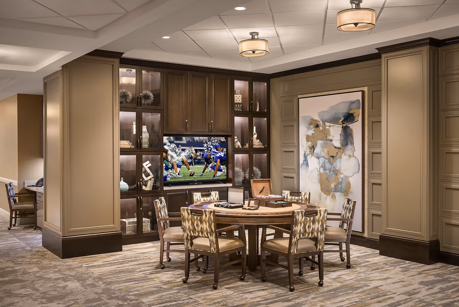A cozy interior room with a round wooden table surrounded by six patterned chairs on casters. The table has a poker set and other game accessories on it. Behind the table is a built-in dark wood cabinet with glass doors displaying decorative items and a flat-screen TV showing a football game. The walls are painted beige with decorative paneling, and there is a large abstract painting on one wall. The ceiling has recessed lighting and two round ceiling lights. The floor is carpeted with a patterned design.