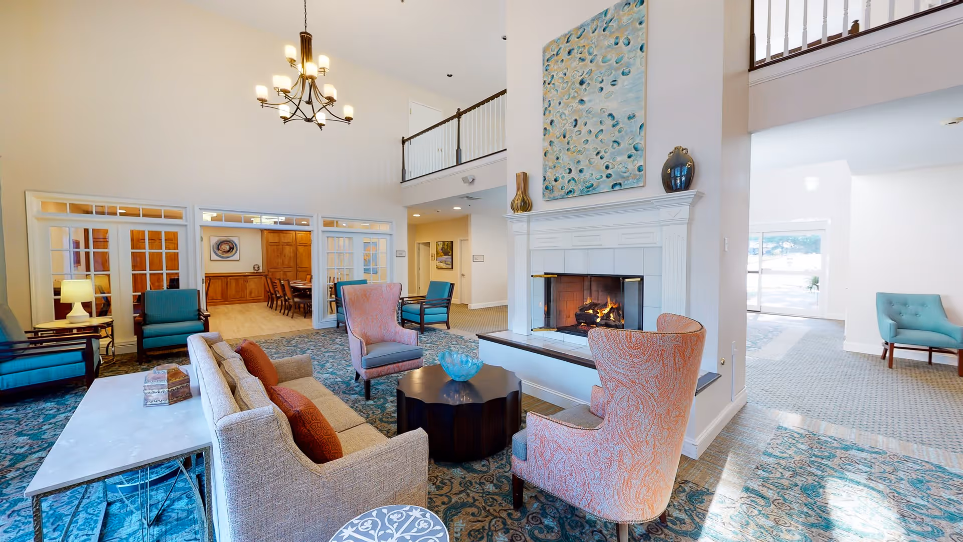 A spacious and well-lit living room area in a senior living facility featuring a fireplace with a large abstract painting above it. The room has a high ceiling with a chandelier, multiple comfortable chairs including two pink patterned armchairs, a beige sofa with orange cushions, and a dark wooden coffee table. The floor is covered with a patterned blue rug, and there is a dining area visible through glass-paneled double doors in the background.