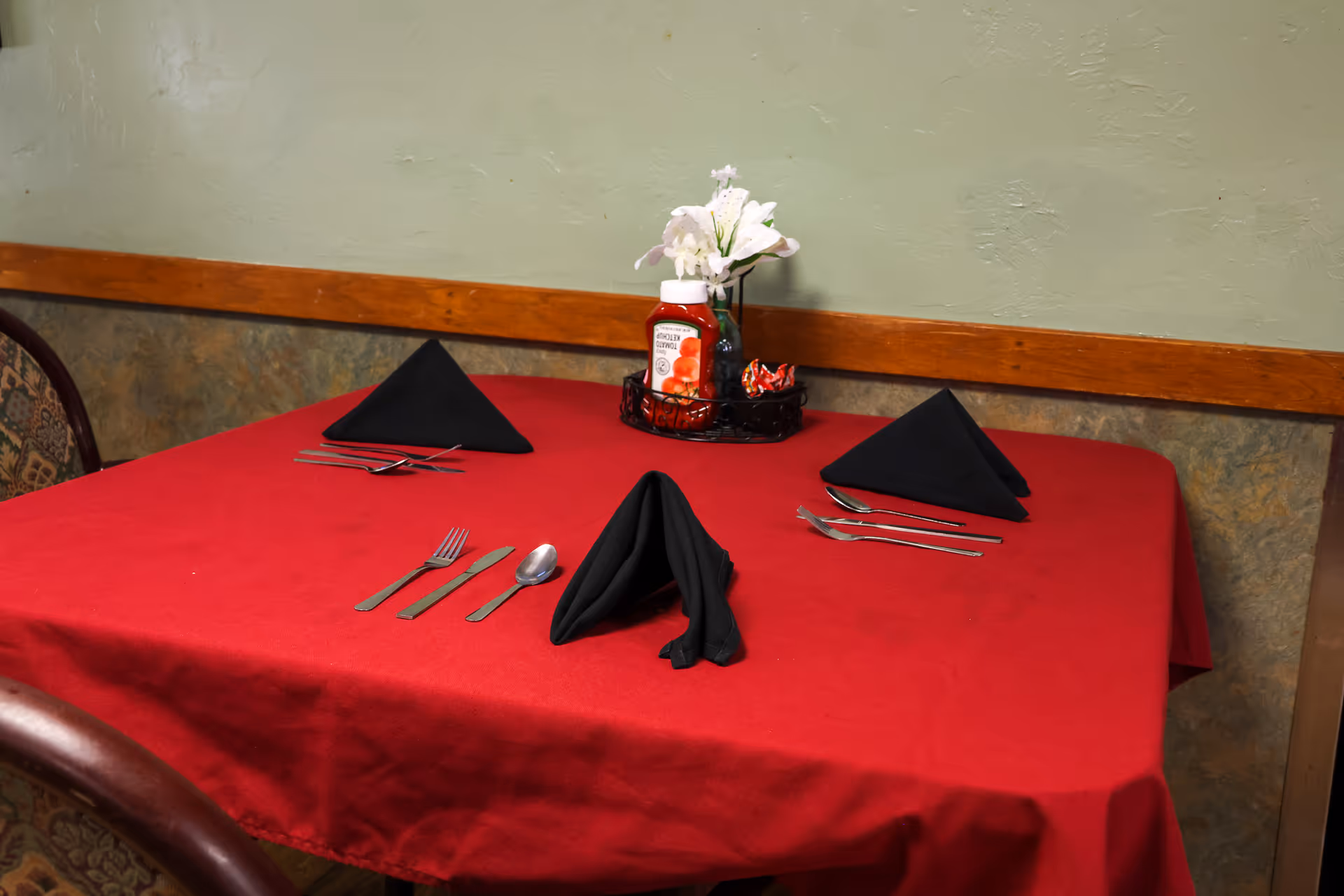 A dining table covered with a red tablecloth set for three people with black folded napkins, silverware, a bottle of ketchup, and a small vase with white flowers against a green wall with wooden trim.