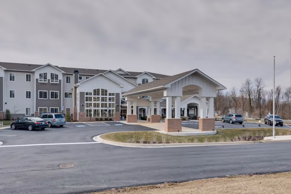 Exterior view of The Landing of Silver Spring senior living facility showing a large three-story building with multiple windows, a covered entrance with white pillars and brick bases, a parking lot with several cars, and a cloudy sky.