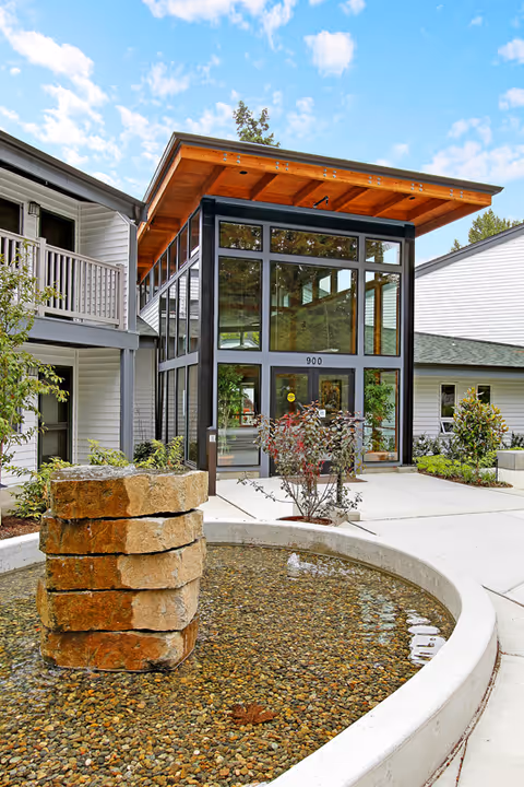 Front entrance of a modern senior living building with large glass doors under a wooden canopy and a circular stone fountain in the foreground.