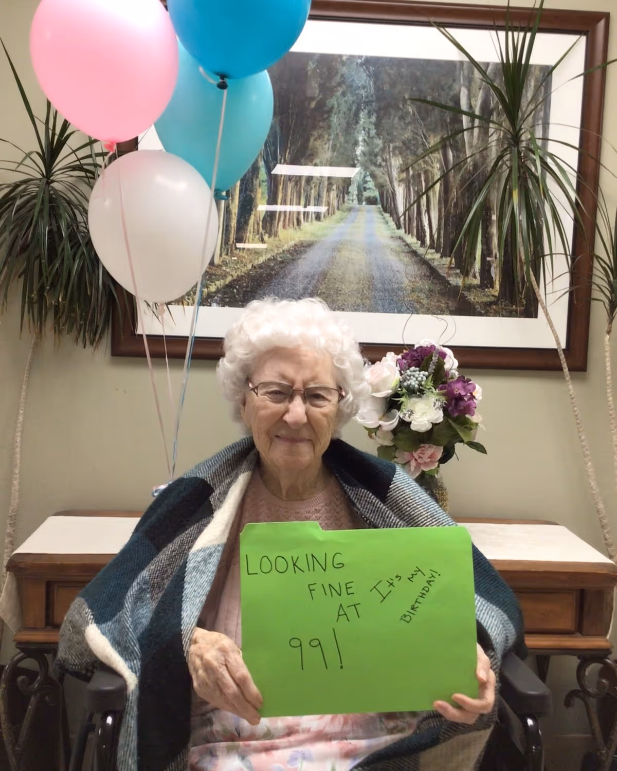 An elderly woman with white curly hair and glasses sits in a wheelchair, wrapped in a plaid blanket. She is holding a green sign that says 'LOOKING FINE AT 99! It's my birthday!' Behind her are colorful balloons, a bouquet of flowers, and a framed picture of a tree-lined path.