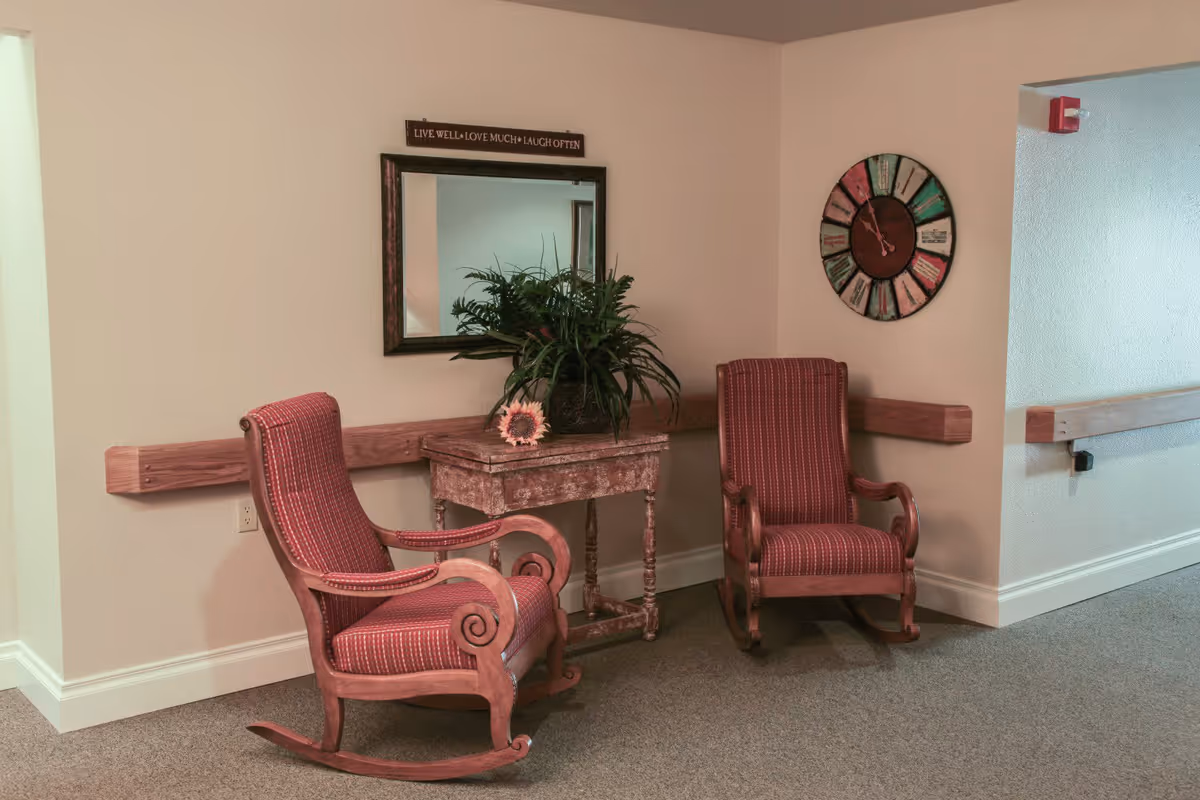 Two red cushioned wooden rocking chairs positioned on either side of a small rustic wooden table with a potted plant and a sunflower decoration. Above the table is a mirror with a sign that reads 'LIVE WELL • LOVE MUCH • LAUGH OFTEN'. A colorful round wall clock is mounted on the adjacent wall. The setting appears to be a quiet hallway or common area with beige walls and carpeted floor.