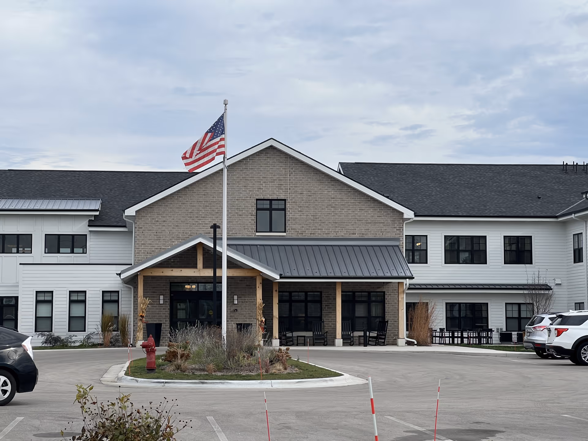 Front exterior view of Lumia Mequon Assisted Living & Memory Care building with a flagpole displaying the American flag, a covered entrance with wooden posts, several windows, parked cars, and a small landscaped area with plants and a fire hydrant.